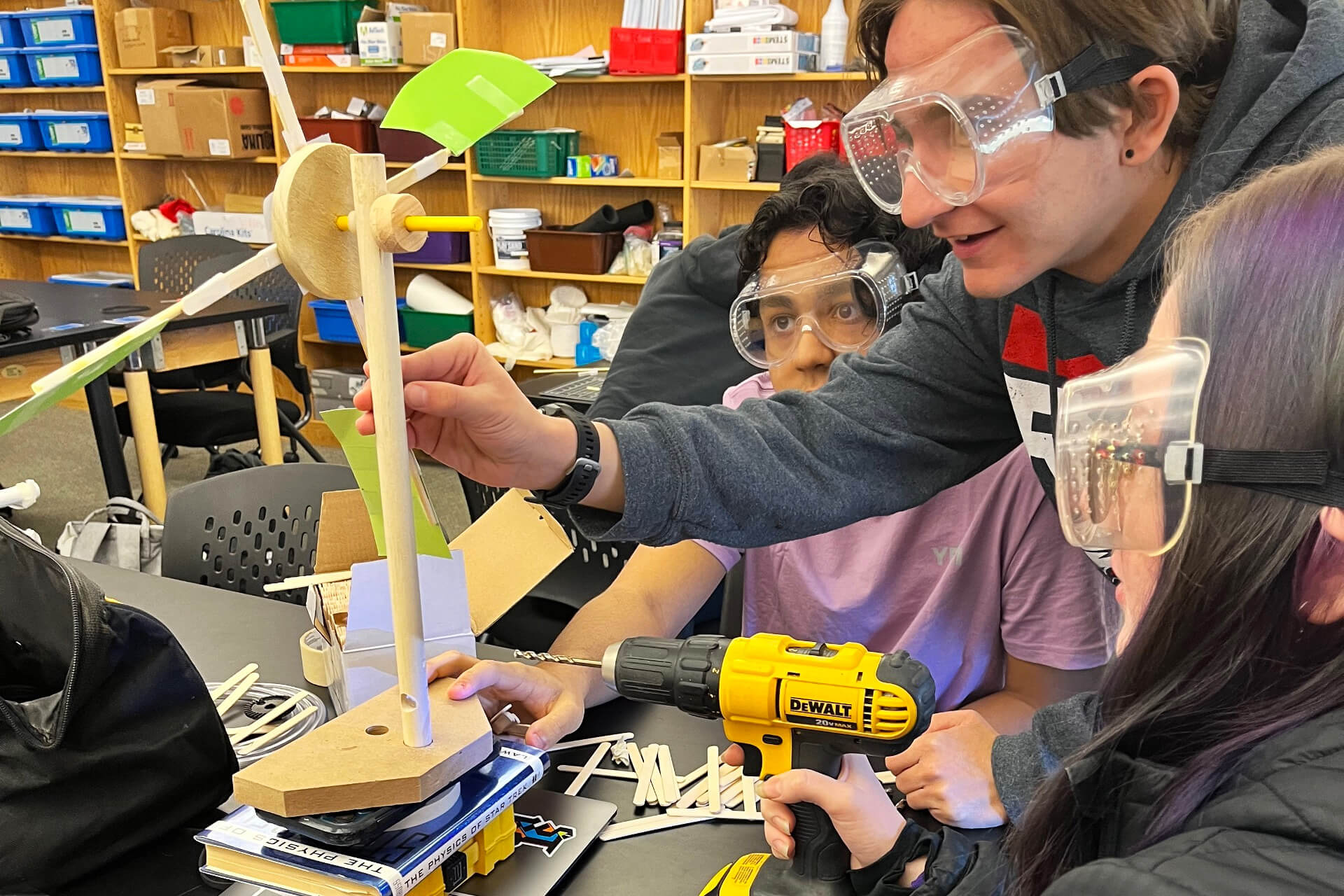A teacher helping students who are working on building a wind indicator using wood and tools.