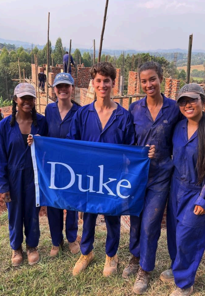 5 students standing next to each other holding Duke University's flag standing on a construction location.