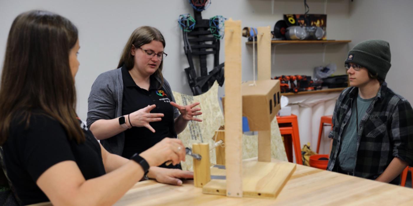 Stacy and Emily and a student around a table and Emily is talking. a prototype of the feeding project on the table.