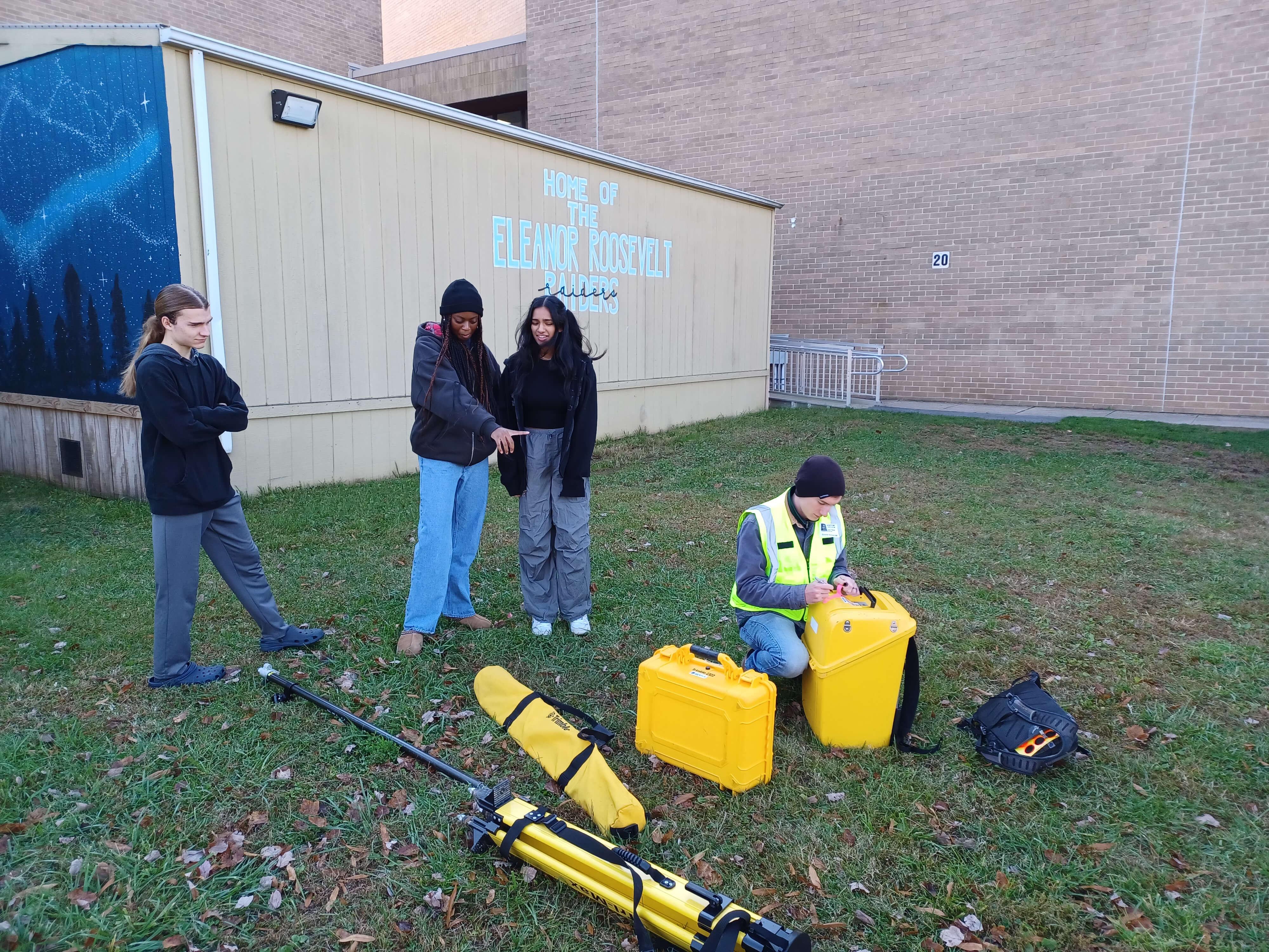 Four students stand outside a school building. Three observe surveying equipment on the ground while one student in a safety vest kneels to prepare bright yellow instrument cases.