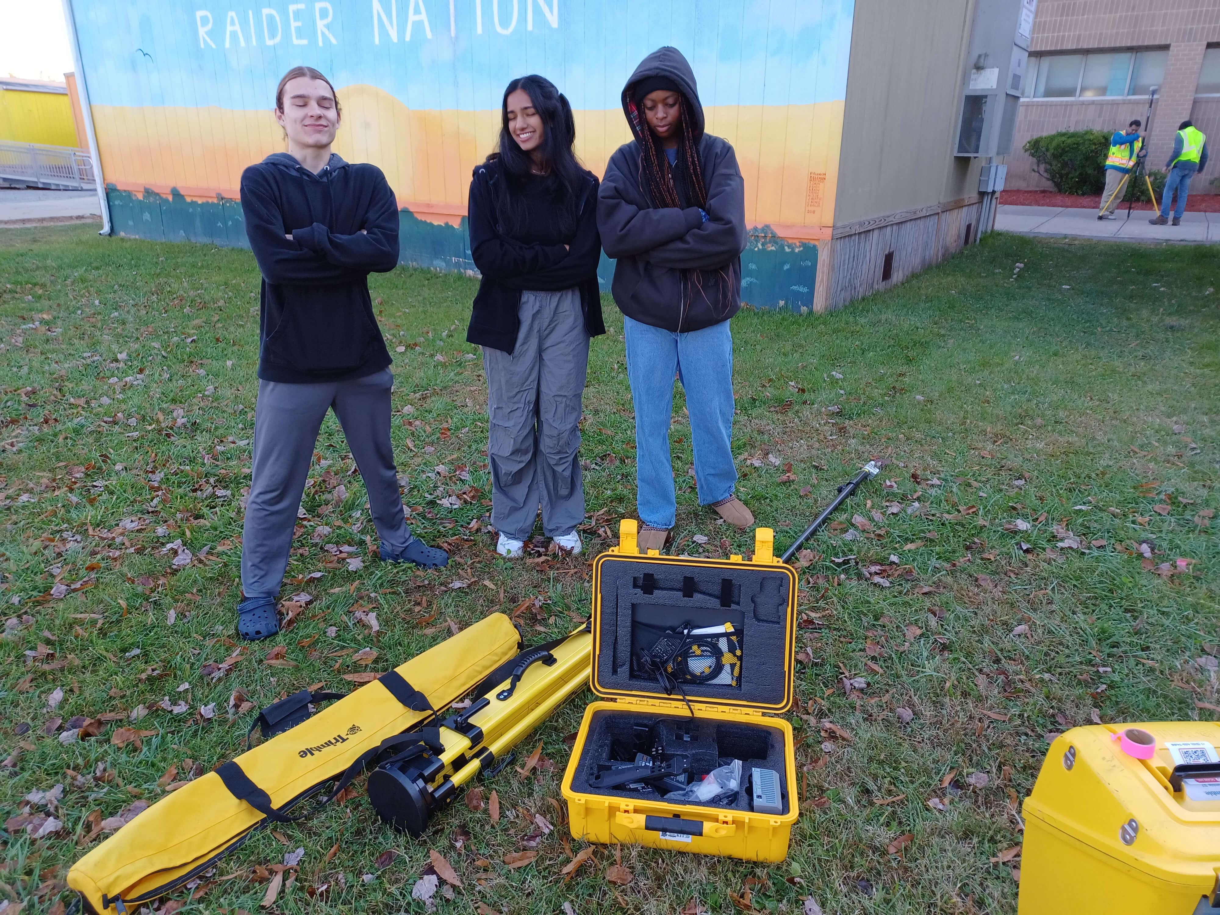 Three students stand smiling with arms crossed near open yellow equipment cases containing surveying tools, while two people in reflective vests work with more surveying equipment in the background.