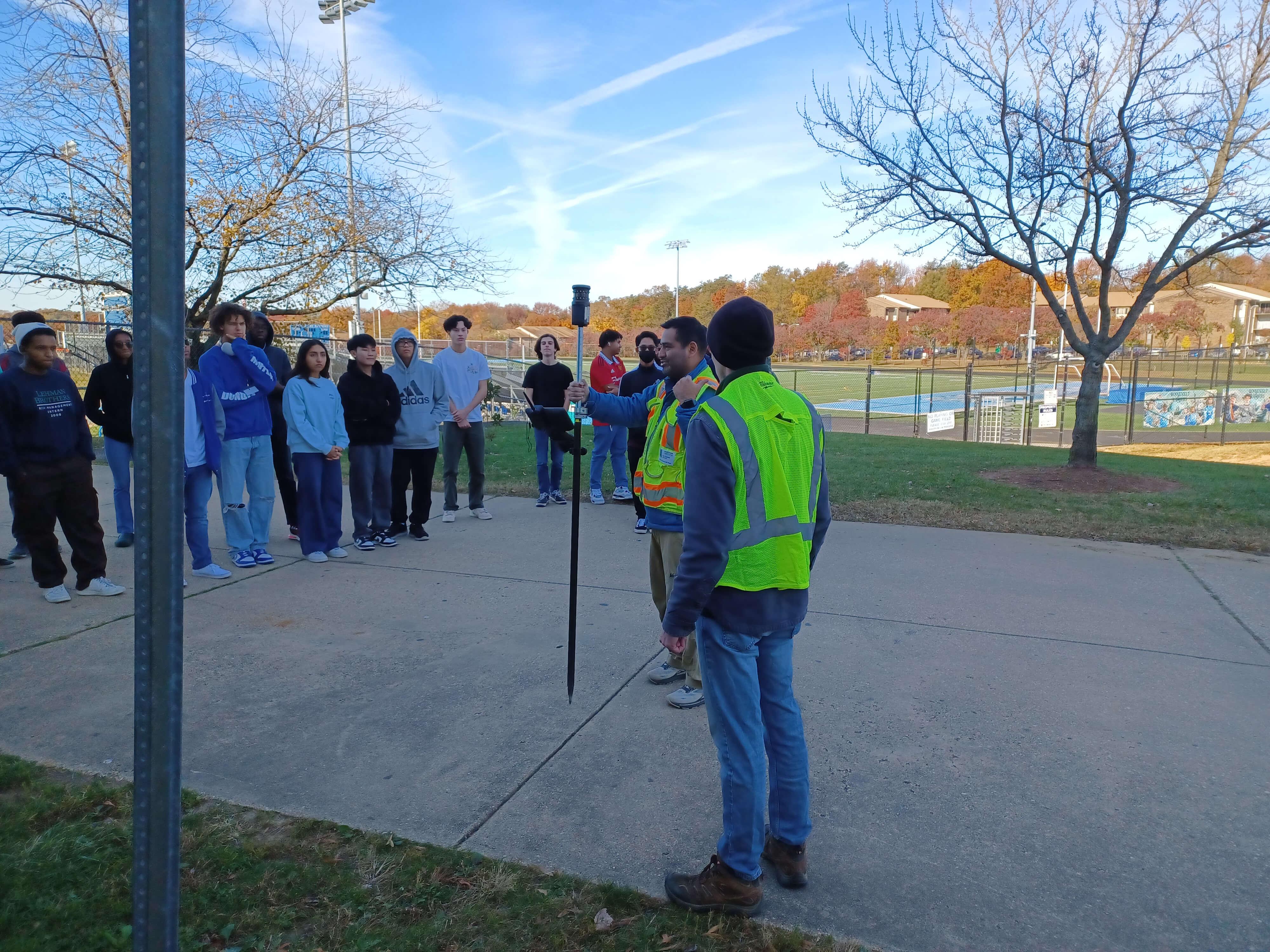 A group of students gathers outdoors as two instructors in safety vests demonstrate how to use a tall surveying instrument near a sports field surrounded by fall-colored trees.