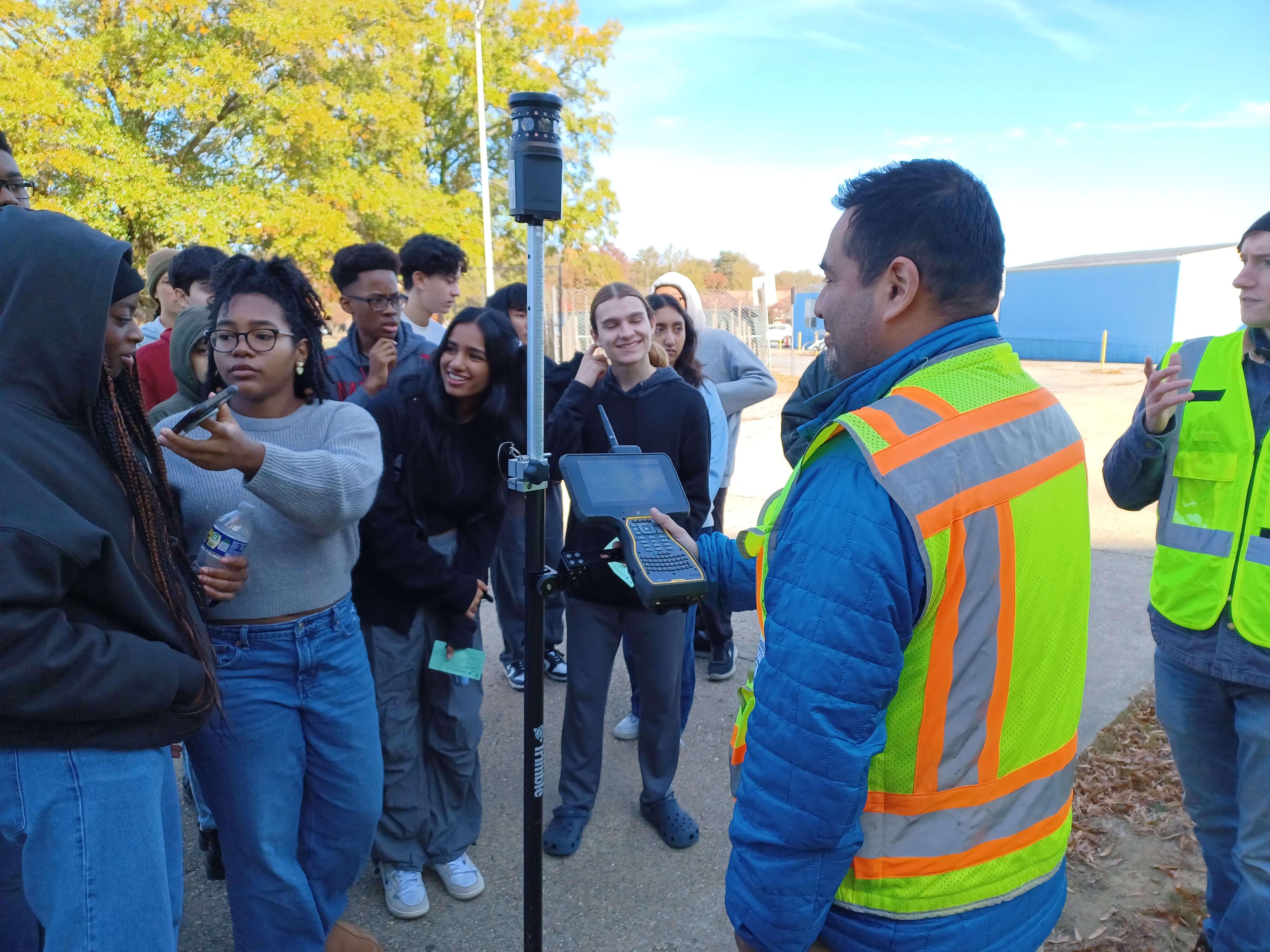 Students crowd around an instructor wearing a bright safety vest who demonstrates a surveying device with a mounted receiver and handheld controller. Several students smile and look closely at the equipment.